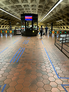 A picture of the faregates and station manager's kiosk in the station. There is blue masking tape on the tile flooring, a rail to the right in the foreground, and a woman walking towards the faregates in the background. The faregates extend to the left and right of the station manager's kiosk in the center, which has a sign on top of it in blue and orange.